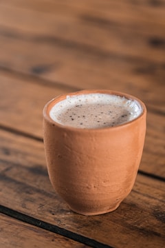 A clay cup filled with frothy liquid is placed on a wooden table with a rustic texture.