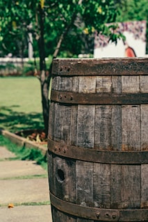 A wooden barrel stands prominently in a lush garden setting. The barrel has metal bands and a rustic, weathered appearance. In the background, there's greenery with trees and bushes, and a path made of stone tiles.