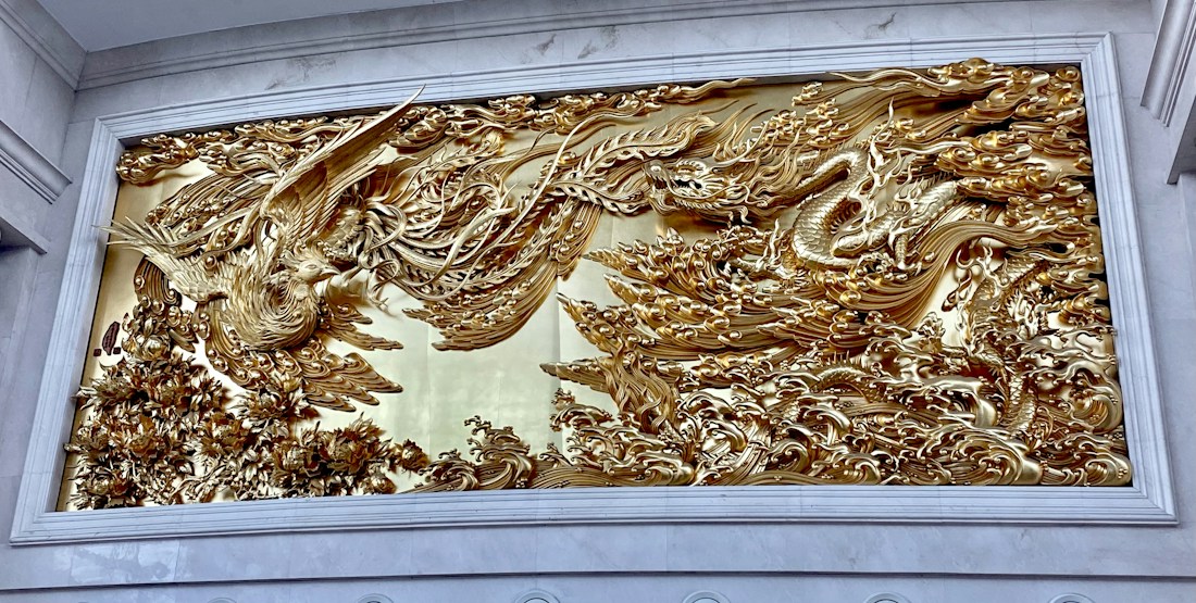 An intricately detailed gold relief sculpture featuring a phoenix on the left and a dragon on the right. The artwork is richly textured with swirling clouds and flames, showcasing the mythological creatures in a dynamic and fluid motion against an ornate backdrop.