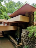 A modern architectural structure with a flat roof, constructed with stone and concrete, surrounded by lush green trees. The building has a series of horizontal lines and terraces that blend with the natural environment. There is a small statue and water feature at the base, adding to the tranquil ambiance.