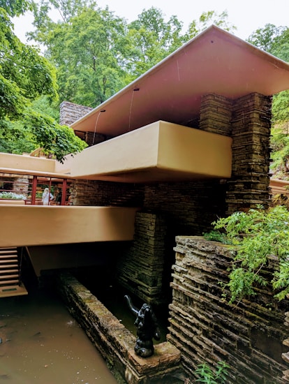 A modern architectural structure with a flat roof, constructed with stone and concrete, surrounded by lush green trees. The building has a series of horizontal lines and terraces that blend with the natural environment. There is a small statue and water feature at the base, adding to the tranquil ambiance.