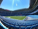 A large, empty football stadium with rows of blue seats surrounding a well-maintained green pitch. The stands are covered with a roof, and a digital screen displays information. The sky is clear and blue, creating a bright and open atmosphere.