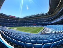 A large, empty football stadium with rows of blue seats surrounding a well-maintained green pitch. The stands are covered with a roof, and a digital screen displays information. The sky is clear and blue, creating a bright and open atmosphere.