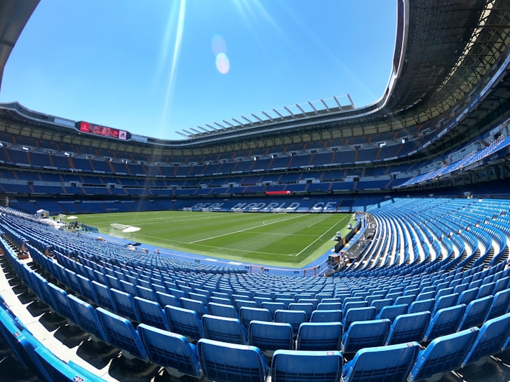 A large, empty football stadium with rows of blue seats surrounding a well-maintained green pitch. The stands are covered with a roof, and a digital screen displays information. The sky is clear and blue, creating a bright and open atmosphere.