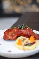 A plated dish featuring a piece of grilled or roasted chicken with a rich red sauce, garnished with chopped chives. Beside the chicken is a serving of creamy mashed potatoes with small carrot pieces and a sprig of herbs. The presentation is elegant and appetizing.