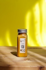 A glass jar filled with turmeric powder is placed on a wooden surface with a bright yellow background. The jar has a metal lid and a label that includes text and a QR code.
