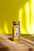 A glass jar filled with turmeric powder is placed on a wooden surface with a bright yellow background. The jar has a metal lid and a label that includes text and a QR code.