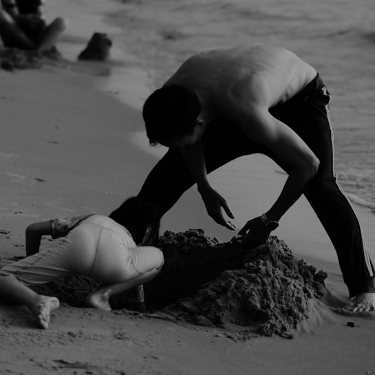 A monochrome photograph captures a person and a child engrossed in building a sand structure by the water's edge. The adult, shirtless and wearing a watch, leans over the child, who is focused on the sand. Their efforts form a small mound, contrasting with the gently lapping waves nearby. The overall scene is relaxed and immersed in the simple joy of beach activities.