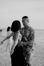 A man and woman are dancing closely on a beach. The man is wearing a floral shirt and shorts, while the woman has a sleeveless dress. The scene has a relaxed and intimate atmosphere, with a blurry background showing a sandy shoreline and another person in the distance.
