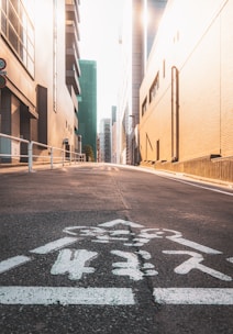 A city street lined with tall buildings on both sides. The asphalt road has a white pedestrian marking with Asian characters. The sun is shining brightly, casting long shadows and creating a warm, golden glow on the buildings.