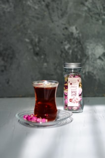 A glass of tea is placed on a clear glass saucer, accompanied by small pink rose buds scattered around. Next to it, a glass jar with a metallic lid contains more rose buds, featuring a decorative label with floral motifs.