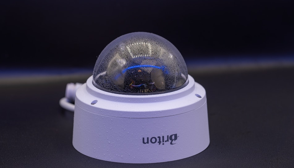 A close-up view of a dome-shaped security camera mounted on a surface. The camera's transparent cover is slightly fogged up. The setup appears to be professional, with visible wiring extending from the back.