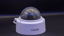 A close-up view of a dome-shaped security camera mounted on a surface. The camera's transparent cover is slightly fogged up. The setup appears to be professional, with visible wiring extending from the back.