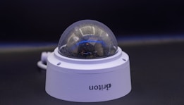 A close-up view of a dome-shaped security camera mounted on a surface. The camera's transparent cover is slightly fogged up. The setup appears to be professional, with visible wiring extending from the back.