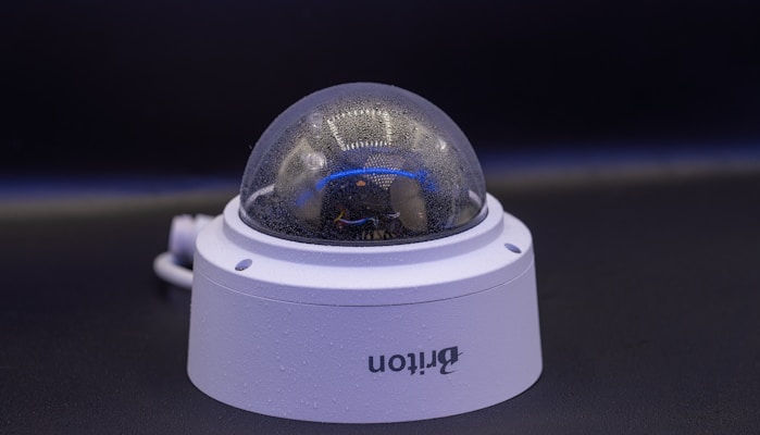 A close-up view of a dome-shaped security camera mounted on a surface. The camera's transparent cover is slightly fogged up. The setup appears to be professional, with visible wiring extending from the back.
