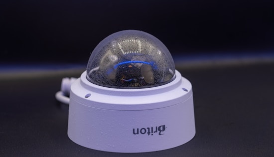 A close-up view of a dome-shaped security camera mounted on a surface. The camera's transparent cover is slightly fogged up. The setup appears to be professional, with visible wiring extending from the back.