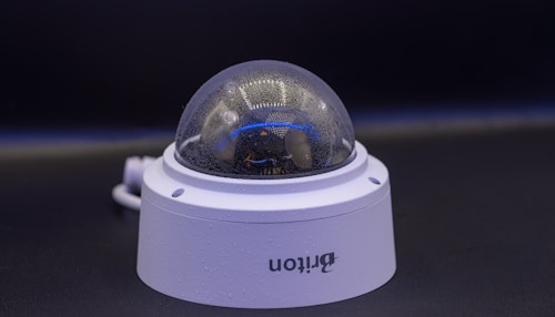 A close-up view of a dome-shaped security camera mounted on a surface. The camera's transparent cover is slightly fogged up. The setup appears to be professional, with visible wiring extending from the back.