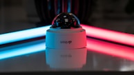 A security camera with a dome-shaped glass cover is placed on a reflective surface. The background features two neon lights, one blue on the left and one red on the right, casting colorful reflections.