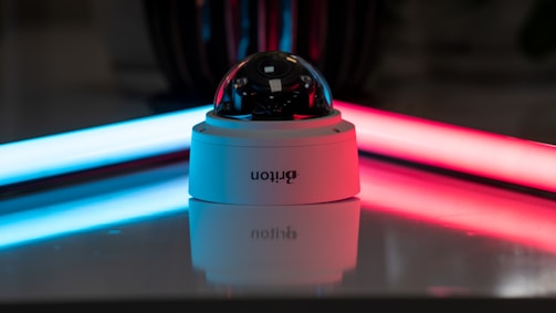 A security camera with a dome-shaped glass cover is placed on a reflective surface. The background features two neon lights, one blue on the left and one red on the right, casting colorful reflections.