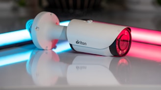 A white security camera is positioned on a reflective surface, with neon lights in blue and pink illuminating the background. The camera has a sleek cylindrical shape with a lens at one end and a mounting bracket at the other.