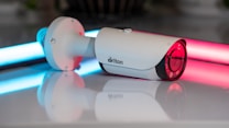 A white security camera is positioned on a reflective surface, with neon lights in blue and pink illuminating the background. The camera has a sleek cylindrical shape with a lens at one end and a mounting bracket at the other.