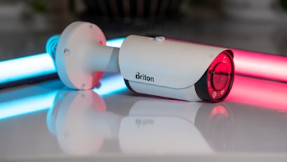 A white security camera is positioned on a reflective surface, with neon lights in blue and pink illuminating the background. The camera has a sleek cylindrical shape with a lens at one end and a mounting bracket at the other.