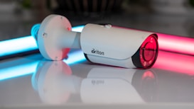 A white security camera is positioned on a reflective surface, with neon lights in blue and pink illuminating the background. The camera has a sleek cylindrical shape with a lens at one end and a mounting bracket at the other.