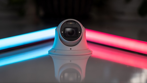 A surveillance camera with a dome shape is positioned on a reflective surface, with two neon lights in the background casting blue and red glows. The camera is centrally placed between the vibrant lights, creating a modern and sleek aesthetic.