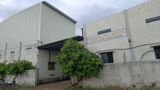 A large industrial building with white and gray walls and a sign that reads 'Tanishq Polymers.' There are green trees in front and a motorcycle parked on the left. The building has small windows and a corrugated metal extension on the roof.