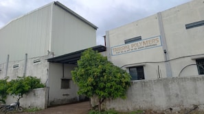 A large industrial building with white and gray walls and a sign that reads 'Tanishq Polymers.' There are green trees in front and a motorcycle parked on the left. The building has small windows and a corrugated metal extension on the roof.