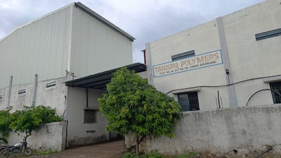 A large industrial building with white and gray walls and a sign that reads 'Tanishq Polymers.' There are green trees in front and a motorcycle parked on the left. The building has small windows and a corrugated metal extension on the roof.