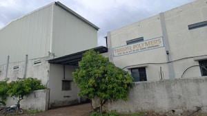 A large industrial building with white and gray walls and a sign that reads 'Tanishq Polymers.' There are green trees in front and a motorcycle parked on the left. The building has small windows and a corrugated metal extension on the roof.