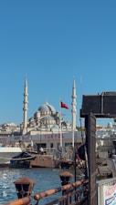 A scenic view of a waterfront area featuring a mosque with prominent domes and minarets in the background. A Turkish flag is visible, and boats are docked by the TURYOL terminal on a sunny day.