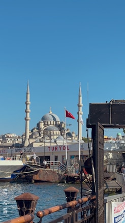 A scenic view of a waterfront area featuring a mosque with prominent domes and minarets in the background. A Turkish flag is visible, and boats are docked by the TURYOL terminal on a sunny day.