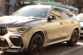 A sleek, modern SUV parked on the side of the street with sunlight reflecting off its polished surface. The vehicle has a sporty design with large alloy wheels and prominent front grill. In the background, there are other cars and urban street elements such as buildings and trees.