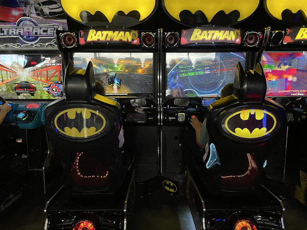 A pair of arcade racing games styled with a Batman theme, featuring distinctive Batman logos on the seats and machine tops. Players engage in a virtual racing experience, with screens displaying vibrant racing graphics and settings.