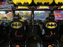 A pair of arcade racing games styled with a Batman theme, featuring distinctive Batman logos on the seats and machine tops. Players engage in a virtual racing experience, with screens displaying vibrant racing graphics and settings.