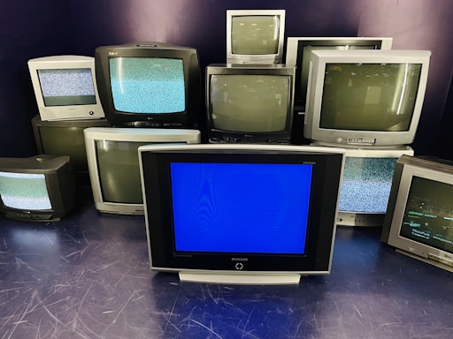 A group of old television sets stacked together with some screens showing static and others off. In the center, one television has a blue screen.