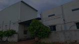 A large industrial building with a white corrugated metal exterior. There is a sign on the facade that reads 'TANISHO POLYMERS' along with an address. The surrounding area includes a fence, some greenery, and a motorcycle parked nearby.
