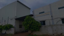 A large industrial building with a white corrugated metal exterior. There is a sign on the facade that reads 'TANISHO POLYMERS' along with an address. The surrounding area includes a fence, some greenery, and a motorcycle parked nearby.