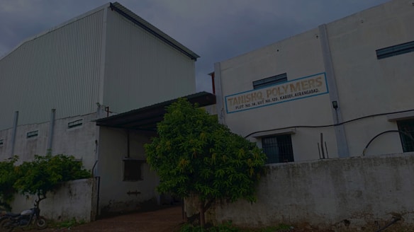 A large industrial building with a white corrugated metal exterior. There is a sign on the facade that reads 'TANISHO POLYMERS' along with an address. The surrounding area includes a fence, some greenery, and a motorcycle parked nearby.