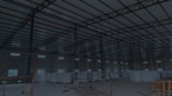 A large, open industrial warehouse interior with a high ceiling and metal beams. Several stacks of large white rolls are placed throughout the area. The space is well-lit by natural light filtering in from rectangular windows along the walls.