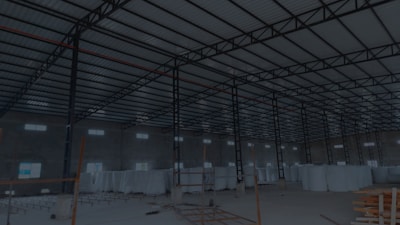 A large, open industrial warehouse interior with a high ceiling and metal beams. Several stacks of large white rolls are placed throughout the area. The space is well-lit by natural light filtering in from rectangular windows along the walls.