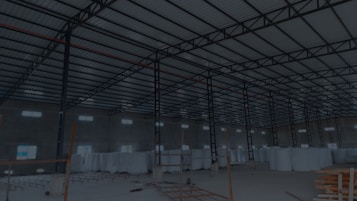 A large, open industrial warehouse interior with a high ceiling and metal beams. Several stacks of large white rolls are placed throughout the area. The space is well-lit by natural light filtering in from rectangular windows along the walls.