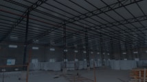 A large, open industrial warehouse interior with a high ceiling and metal beams. Several stacks of large white rolls are placed throughout the area. The space is well-lit by natural light filtering in from rectangular windows along the walls.
