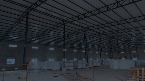 A large, open industrial warehouse interior with a high ceiling and metal beams. Several stacks of large white rolls are placed throughout the area. The space is well-lit by natural light filtering in from rectangular windows along the walls.