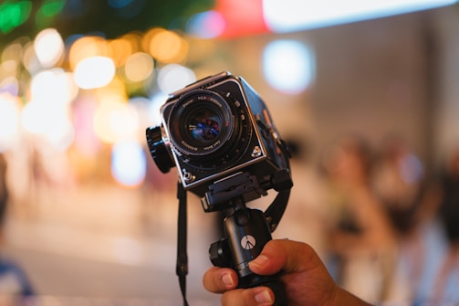 A hand holds a vintage camera with a lens facing forward, set against a blurred background with bokeh lights, creating a vibrant and colorful atmosphere.