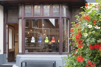 A boutique store facade featuring a window display with colorful bust sculptures and eyeglasses. The storefront is framed by ornate dark wood and stained glass, with a vibrant arrangement of red and white flowers on the right.