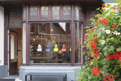A boutique store facade featuring a window display with colorful bust sculptures and eyeglasses. The storefront is framed by ornate dark wood and stained glass, with a vibrant arrangement of red and white flowers on the right.