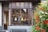 A boutique store facade featuring a window display with colorful bust sculptures and eyeglasses. The storefront is framed by ornate dark wood and stained glass, with a vibrant arrangement of red and white flowers on the right.